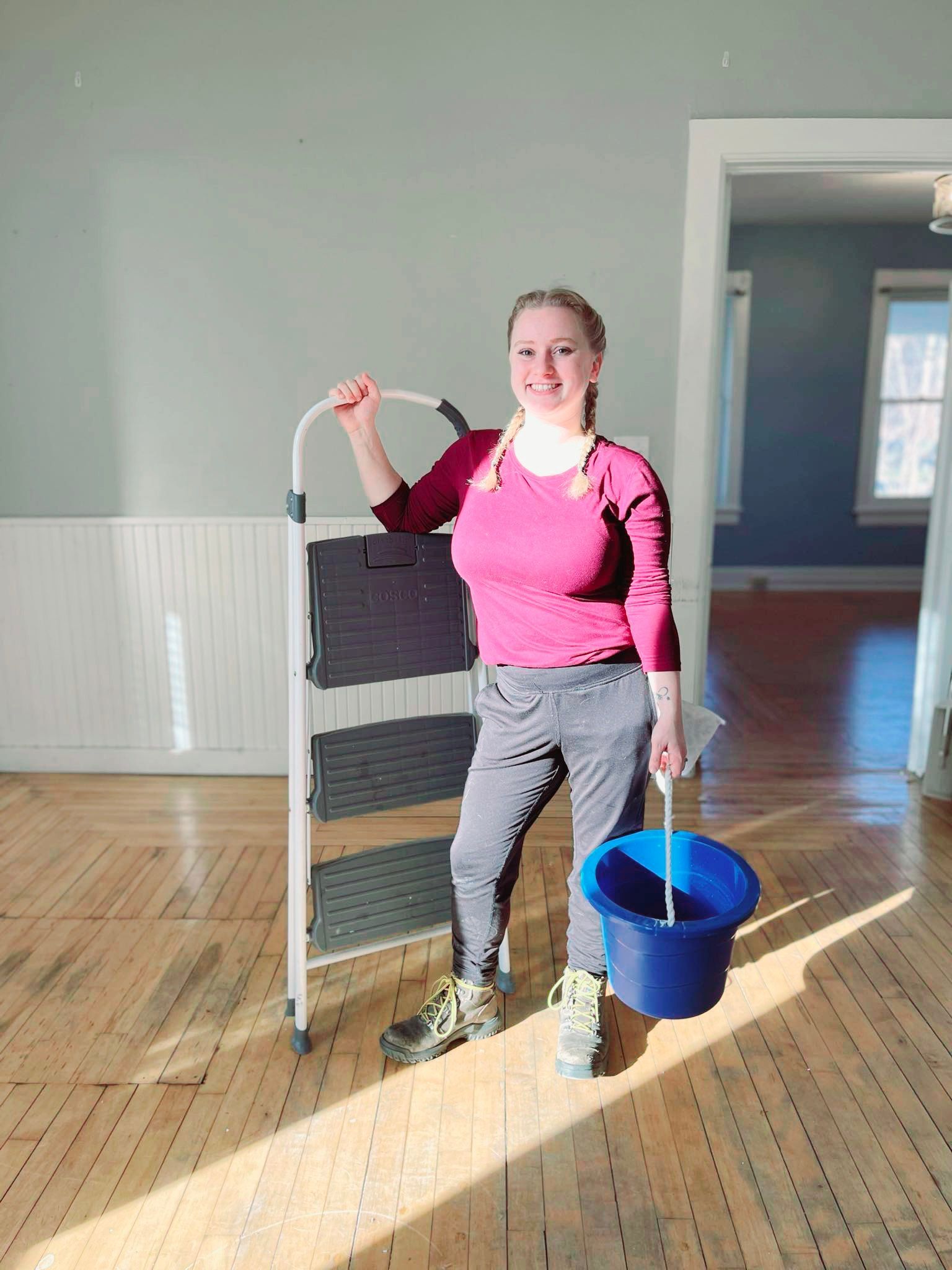 Woman with ladder, bucket, in room with wood floor and light-blue walls.