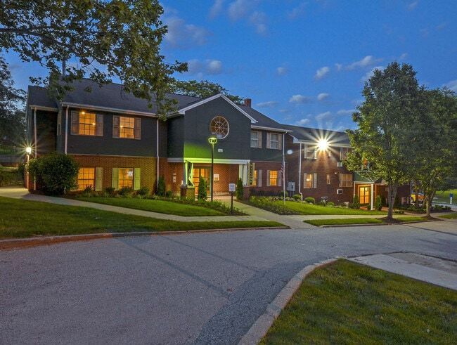 Multi-story brick building with green trim, lit at dusk. Landscaped grounds, street in foreground.
