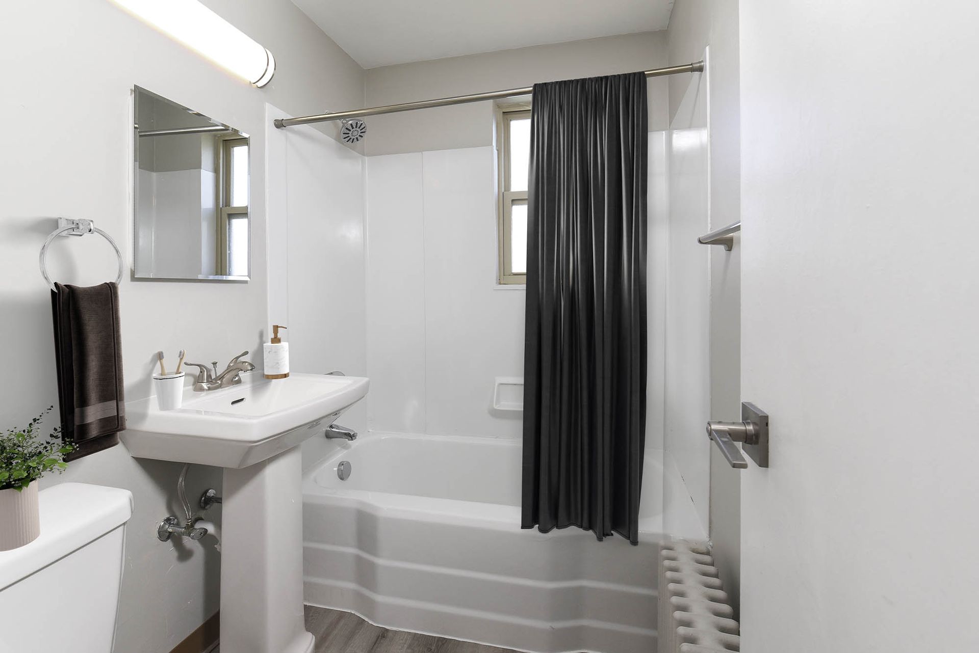 Small white apartment bathroom with pedestal sink, tub/shower, mirror, and a dark shower curtain at The Alden South Hills in Pittsburgh, PA. 