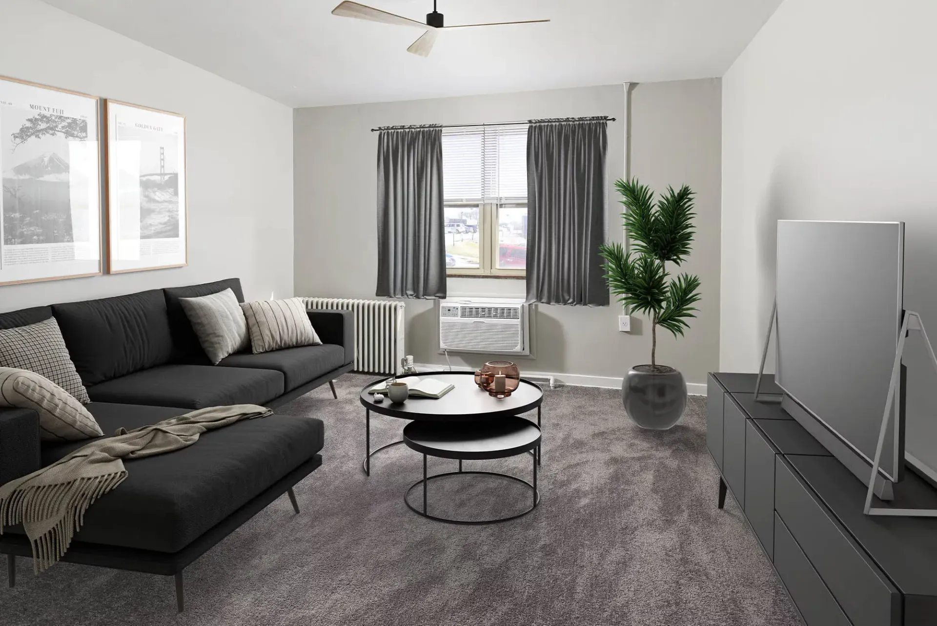Living room in an apartment with a dark gray sofa, coffee table, curtains, TV stand, and a plant at The Alden South Hills in Pittsburgh, PA. 