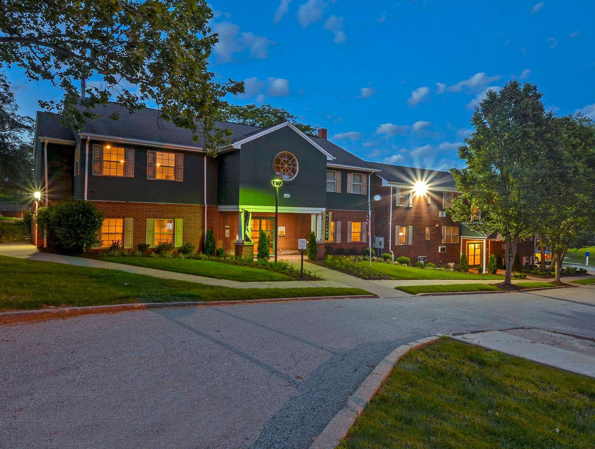 Exterior view of a two-story apartment complex at dusk with lit windows and landscaped grounds at The Alden South Hills in Pittsburgh, PA. 