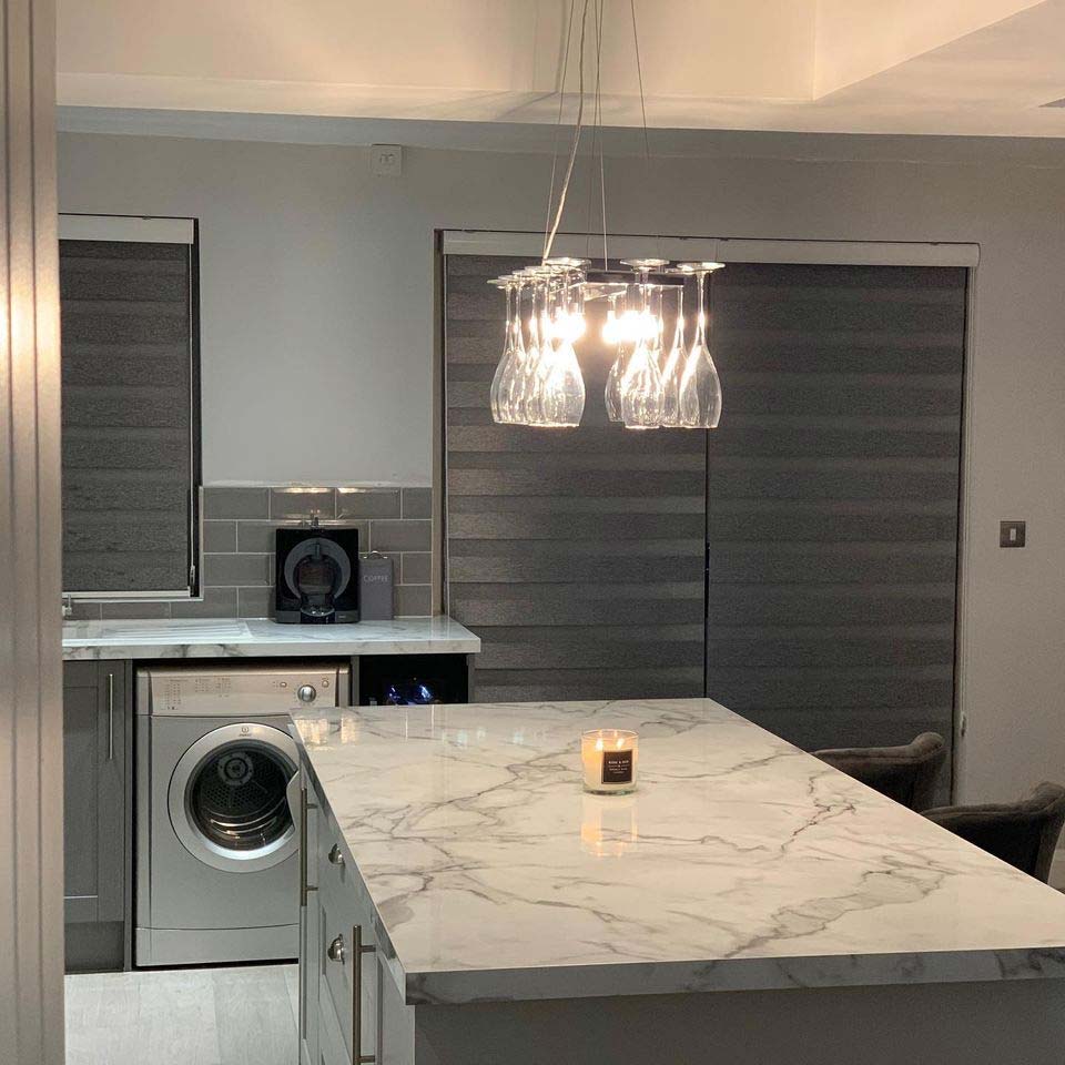 A modern kitchen with a marble island and low hanging light fixture with sleek grey blinds