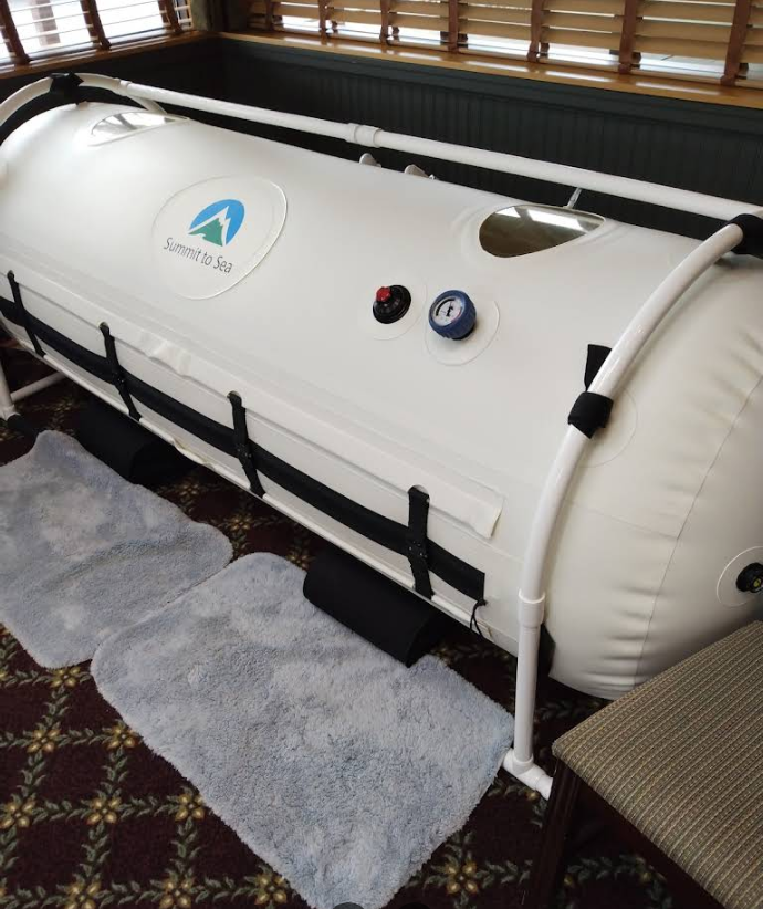 White hyperbaric chamber with black straps and a blue logo. Resting on a white frame, with two blue foot mats.