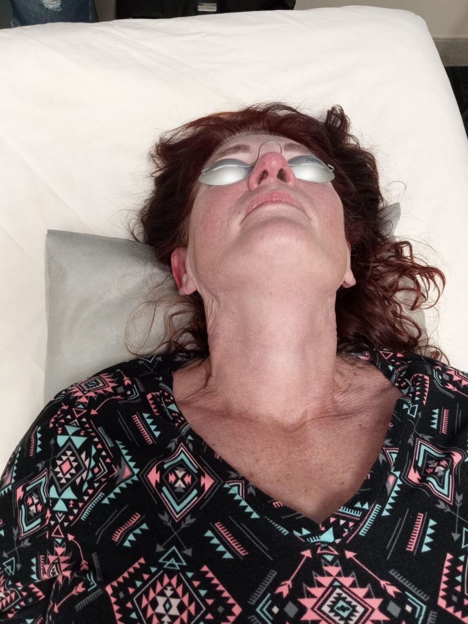 Person with protective eyewear, reclined on a bed, looking upwards. Neck and chest are visible, wearing a patterned top.