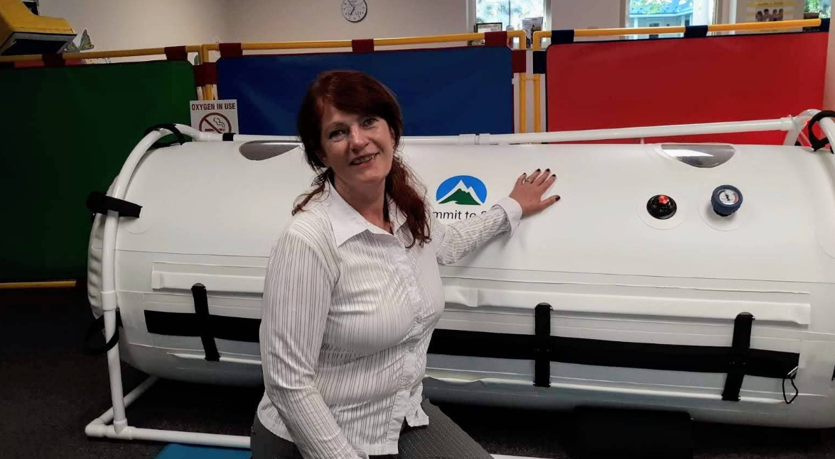Woman points to a white hyperbaric chamber with gauges, colorful panels in the background.