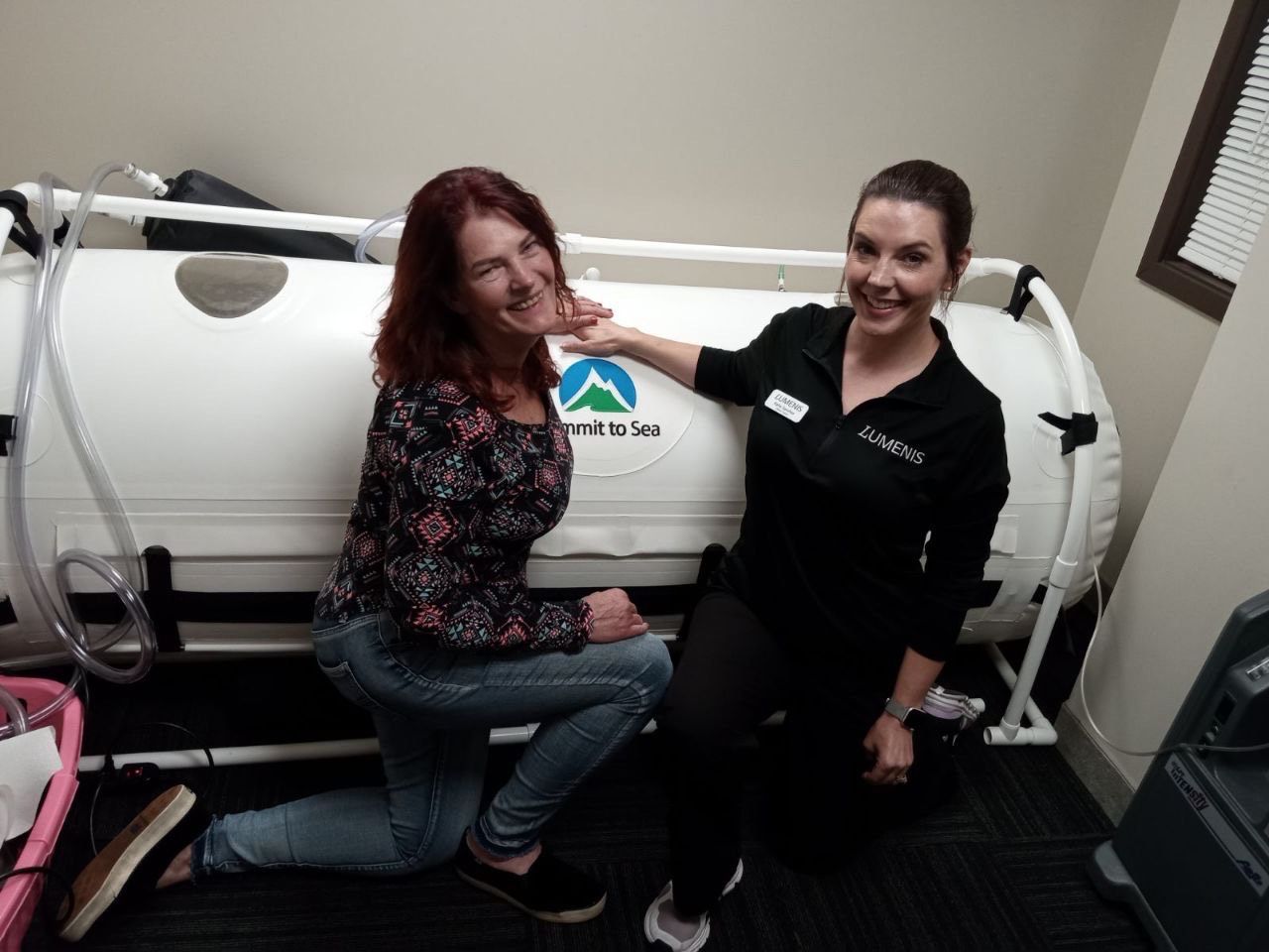 Two women posing by a hyperbaric oxygen therapy chamber; one points to the chamber, both smile.
