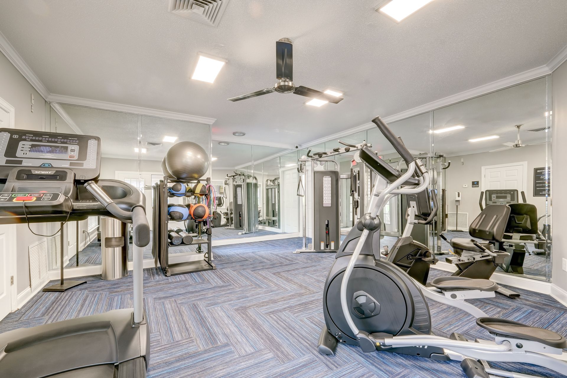 A brightly lit gym with a blue carpet, featuring a treadmill, an elliptical machine, and a rack of fitness balls.