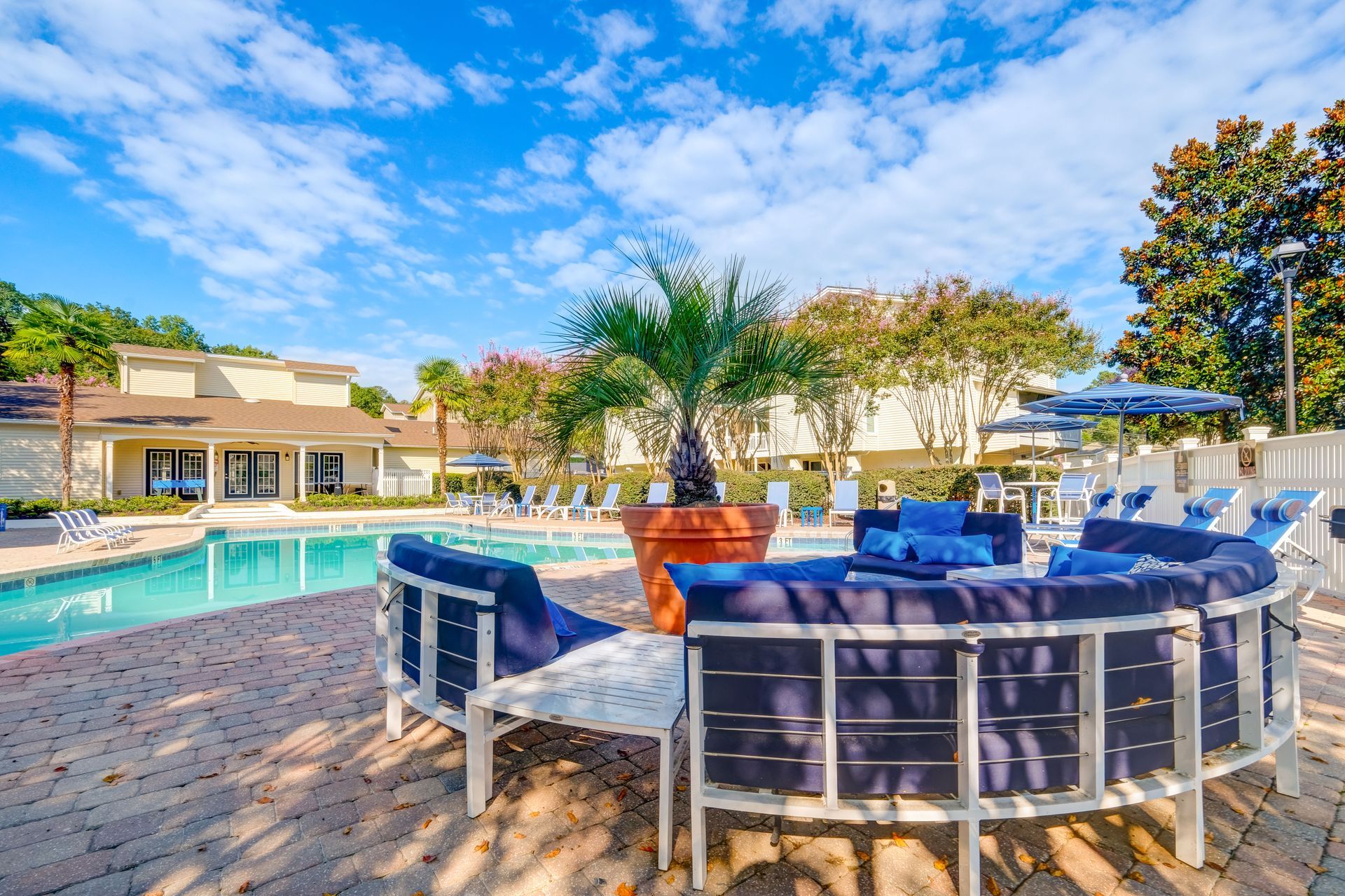 A semicircular outdoor lounge area with dark blue cushions sits on a brick patio by a swimming pool under a sunny sky.