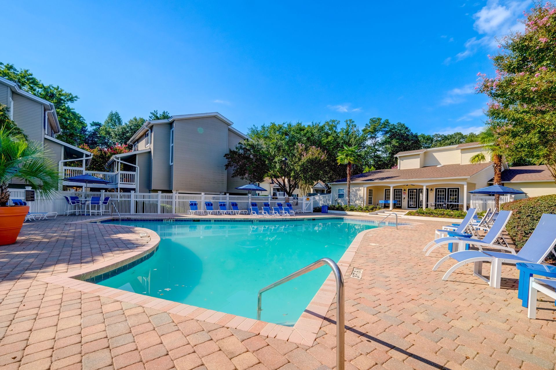 A sunny swimming pool surrounded by a brick patio, lounge chairs, and apartment buildings under a clear blue sky.