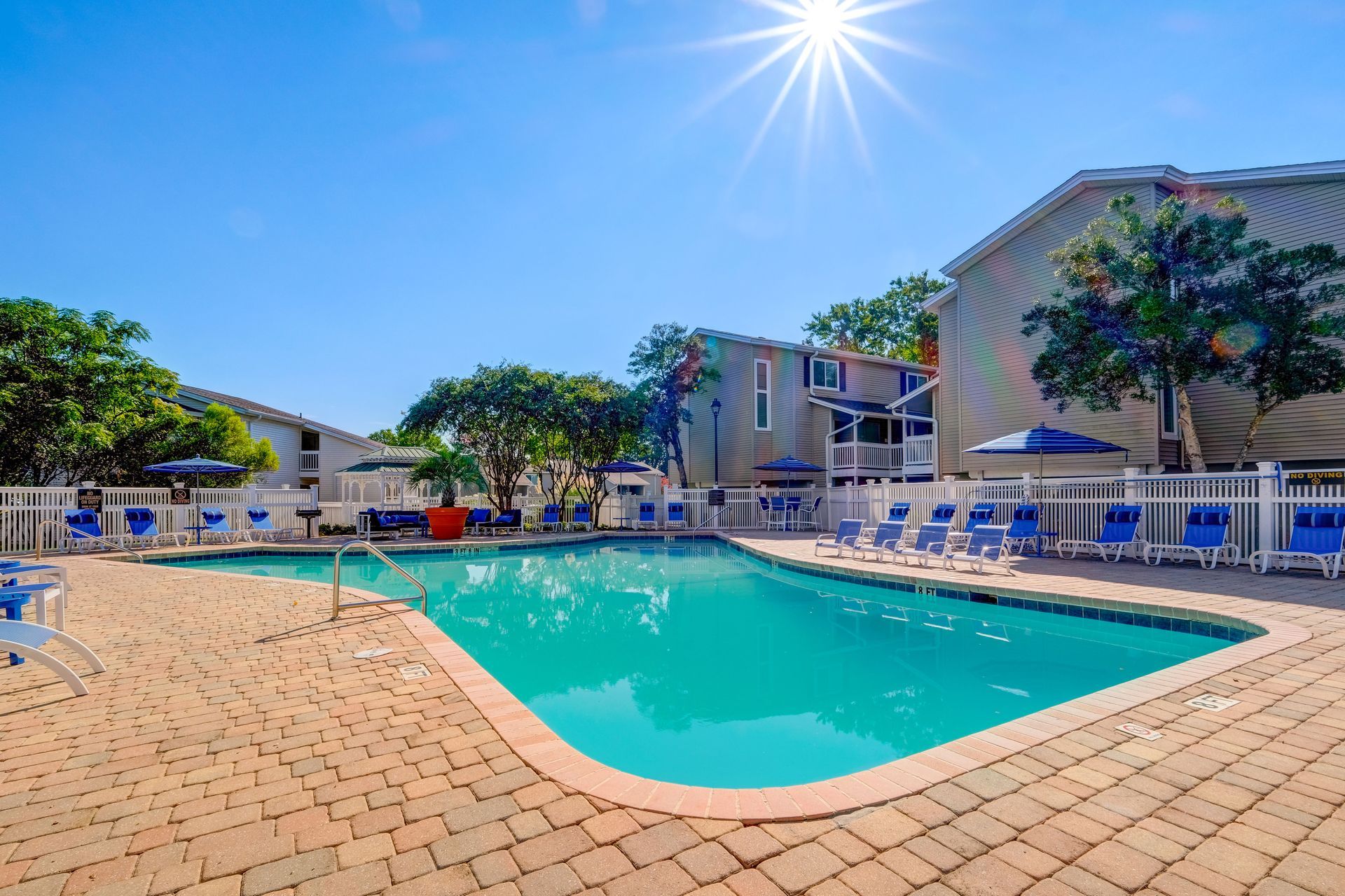 A sunny outdoor swimming pool surrounded by a brick patio, lounge chairs, and multistory apartment buildings.