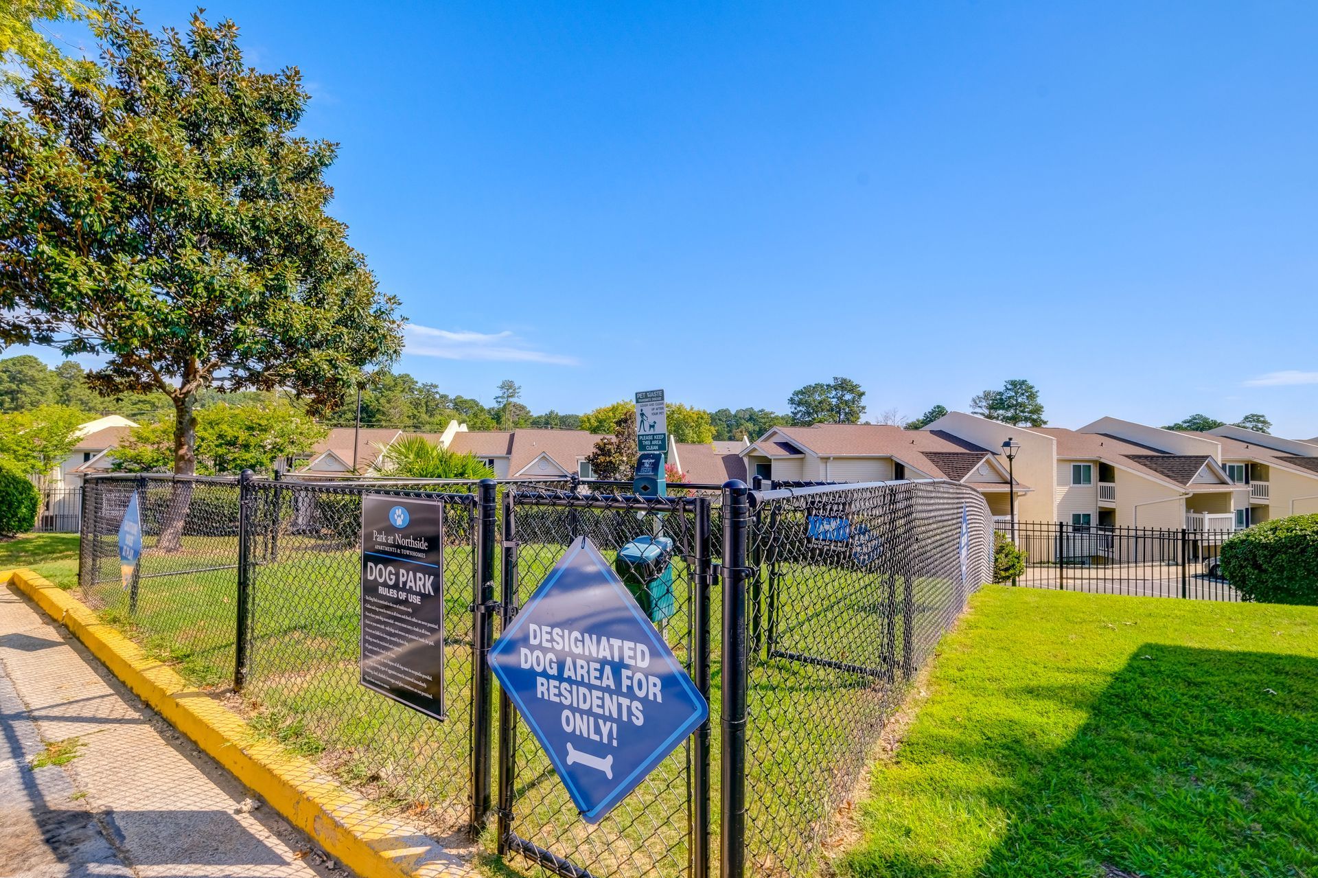 A fenced-in dog park area features a blue sign with white text, lush green grass, a large tree, and apartments in the back.