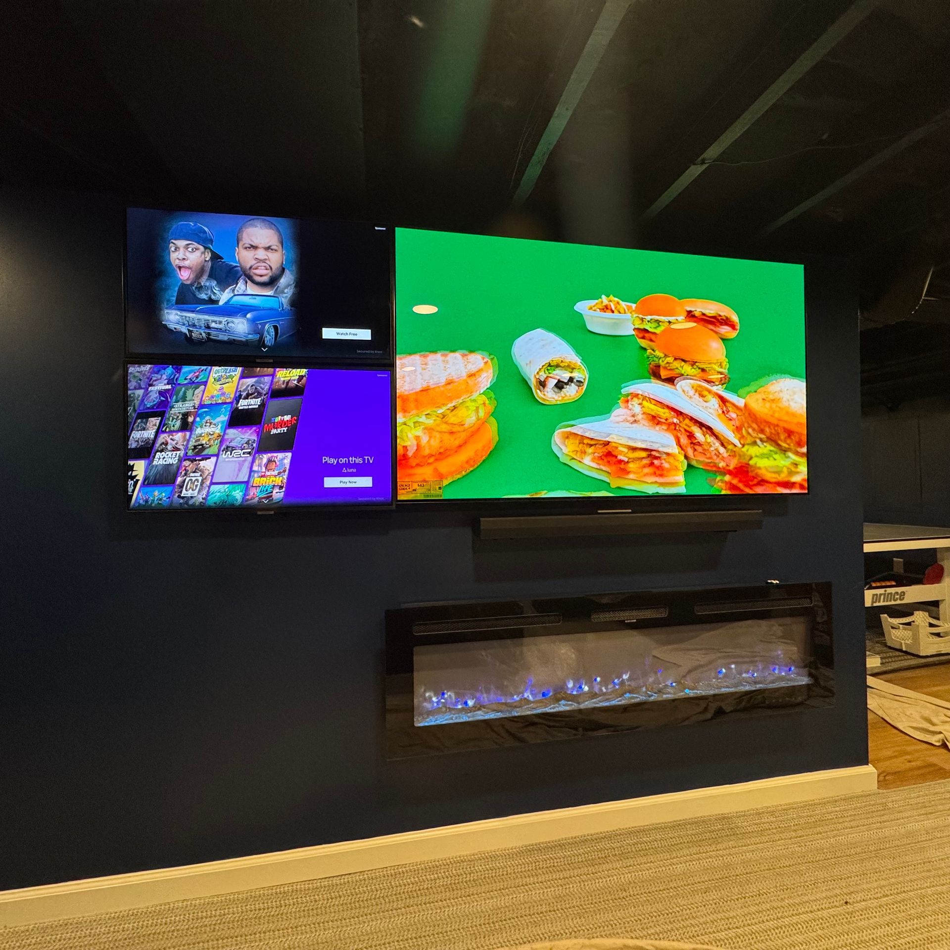 Four TVs mounted on a dark blue wall above a fireplace. One TV shows two men, one shows food, and one shows a streaming service.