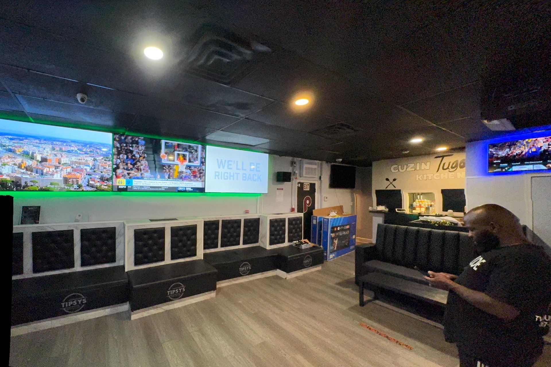 Interior of a lounge with multiple screens showing images. Black couches and seating line the walls, and a man stands in the foreground.