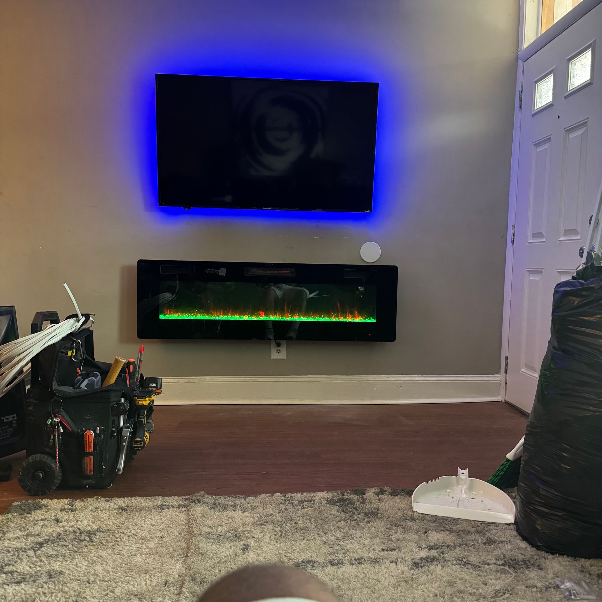 A mounted TV with blue backlighting sits above a wall-mounted electric fireplace. An open door and cleaning supplies are nearby.