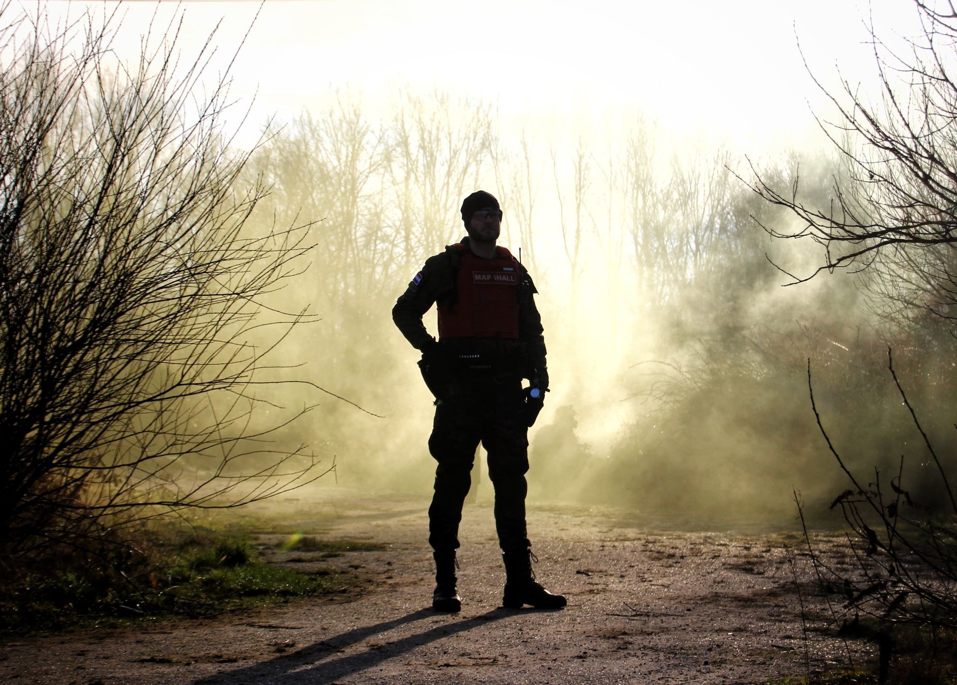 A person in tactical gear stands on a path, silhouetted against smoke and trees.