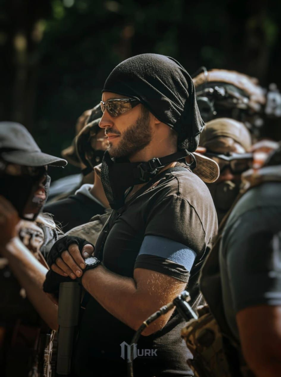 Man in black beanie and tactical gear, smiling. Outdoors, surrounded by others in similar attire.