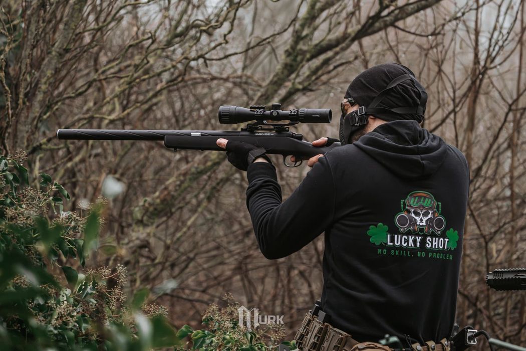 Person aiming a rifle with scope in a wooded outdoor setting, wearing a black hoodie.
