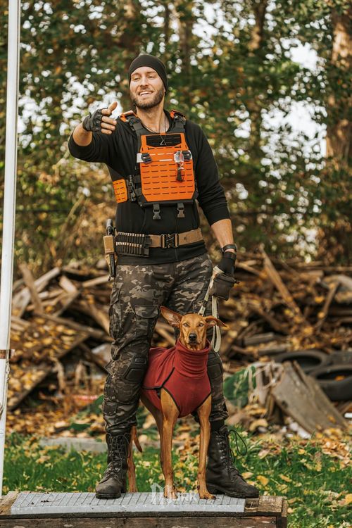 Man in orange vest standing outdoors with a dog in a red harness, holding up a small object.