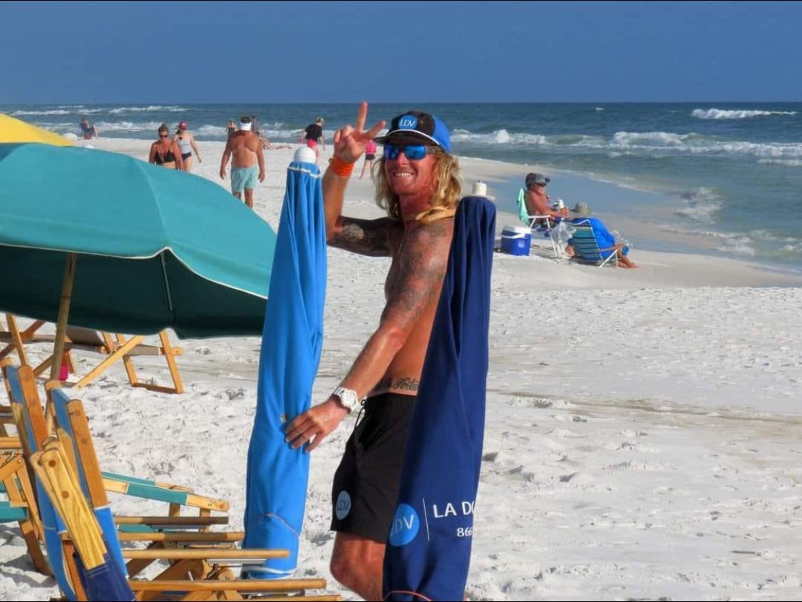 A shirtless man is standing on a beach next to a blue umbrella