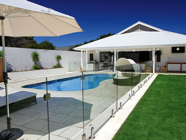 Modern backyard pool with glass fence, white patio, lounge chairs, and umbrella under a blue sky