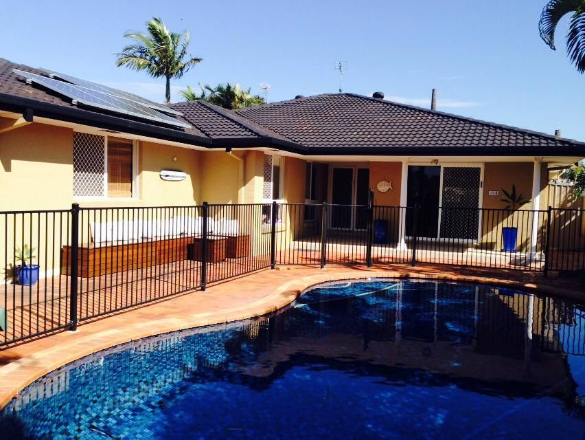 Backyard patio with a blue swimming pool, black fence, and a single-story house under a sunny sky