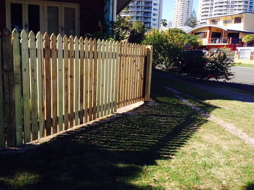 Wooden picket fence beside a house, casting shadows on a grassy yard near a street.