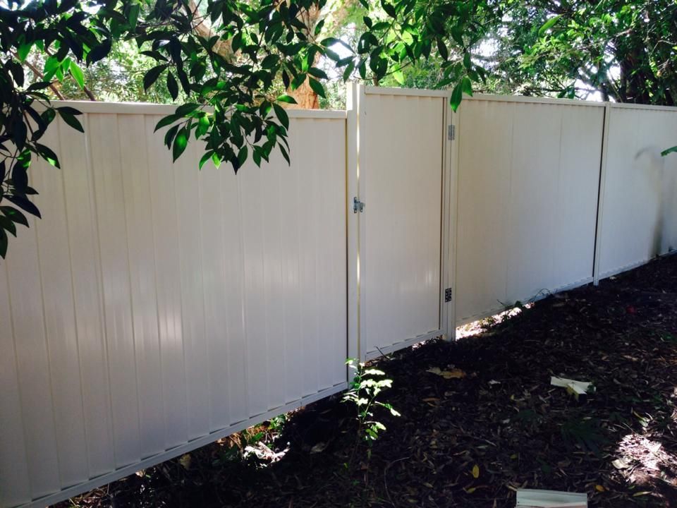White vinyl fence along a shaded dirt path with tree branches overhead and plants at the base
