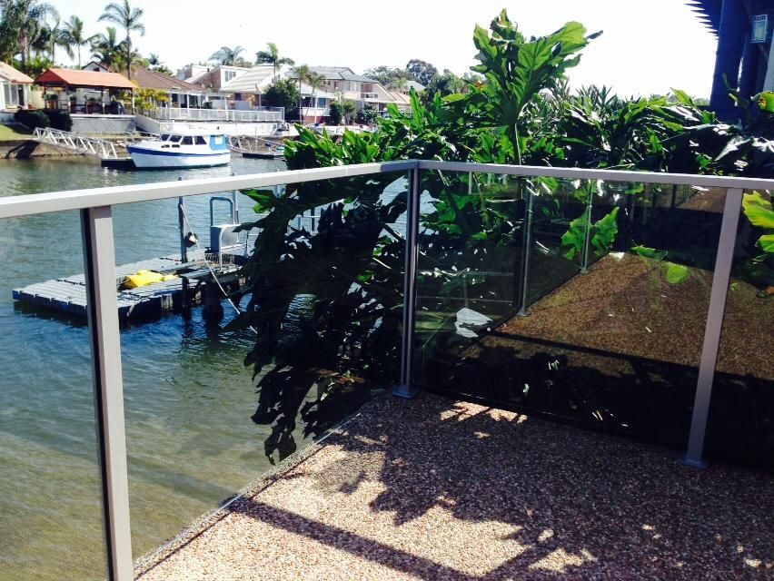 Balcony with glass railing overlooking a canal, boats, and tropical plants in bright sunlight