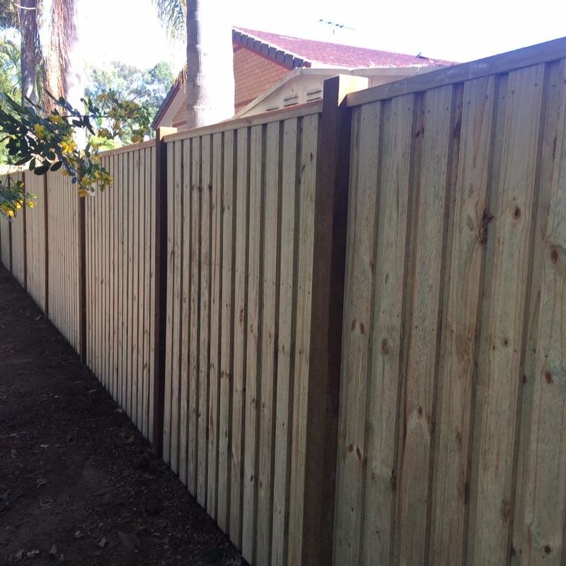 Tall wooden privacy fence along a narrow dirt path beside a house and trees.