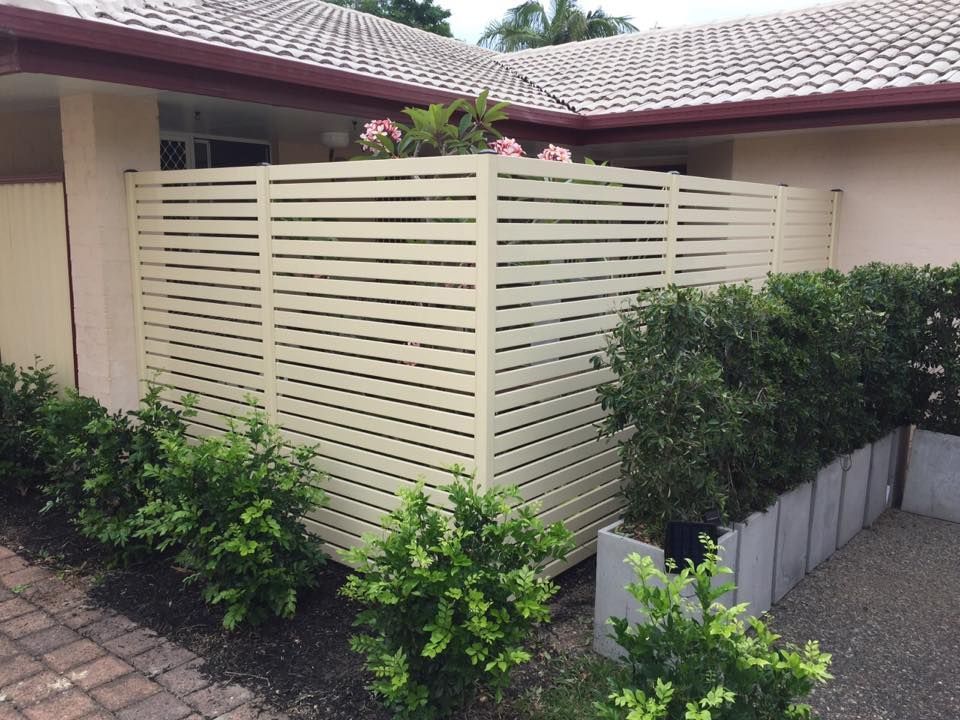 Cream slatted fence beside a house, bordered by shrubs and a paved driveway.