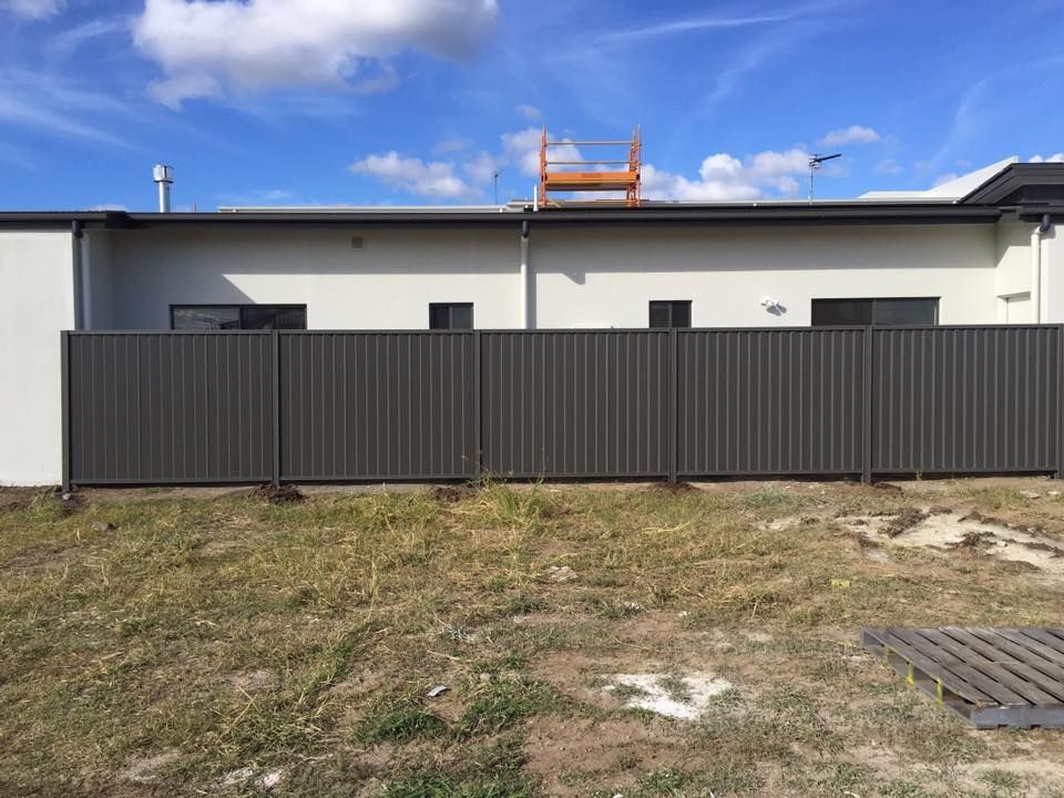 Modern house behind a dark fence under a blue sky, with a patchy grassy foreground.