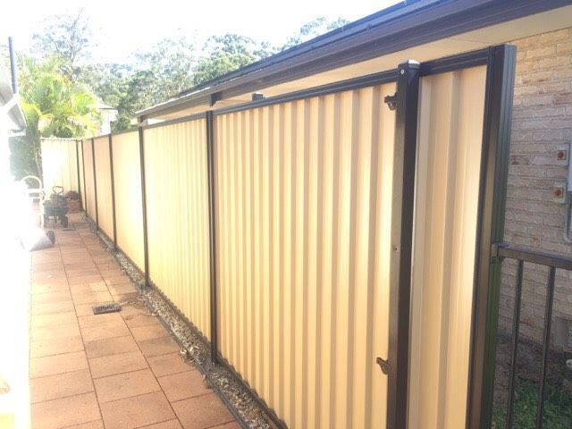 Long beige corrugated metal fence along a brick walkway beside a house.