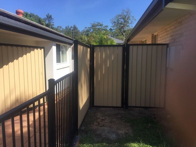Narrow outdoor passage between houses with tan fencing, black railing, and a gate in bright sunlight