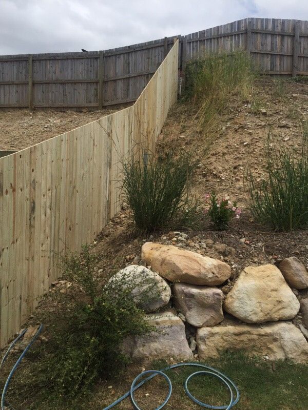 Wooden fence on a sloped dirt yard with rocks, weeds, and a coiled blue hose near a retaining wall
