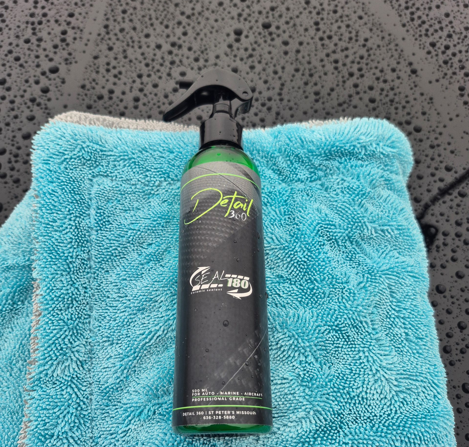 A green detailing spray bottle on a blue microfiber towel, against a wet, dark car surface.