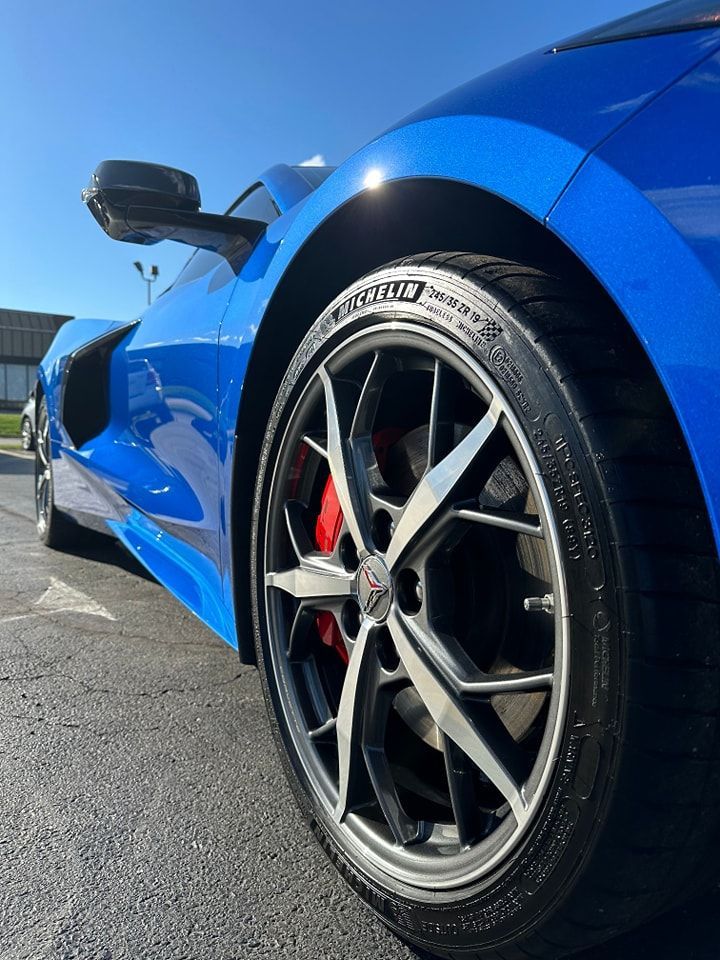 Blue sports car, close-up of a front wheel. Red brake caliper, silver and black rim, Michelin tire. Bright sunny day.
