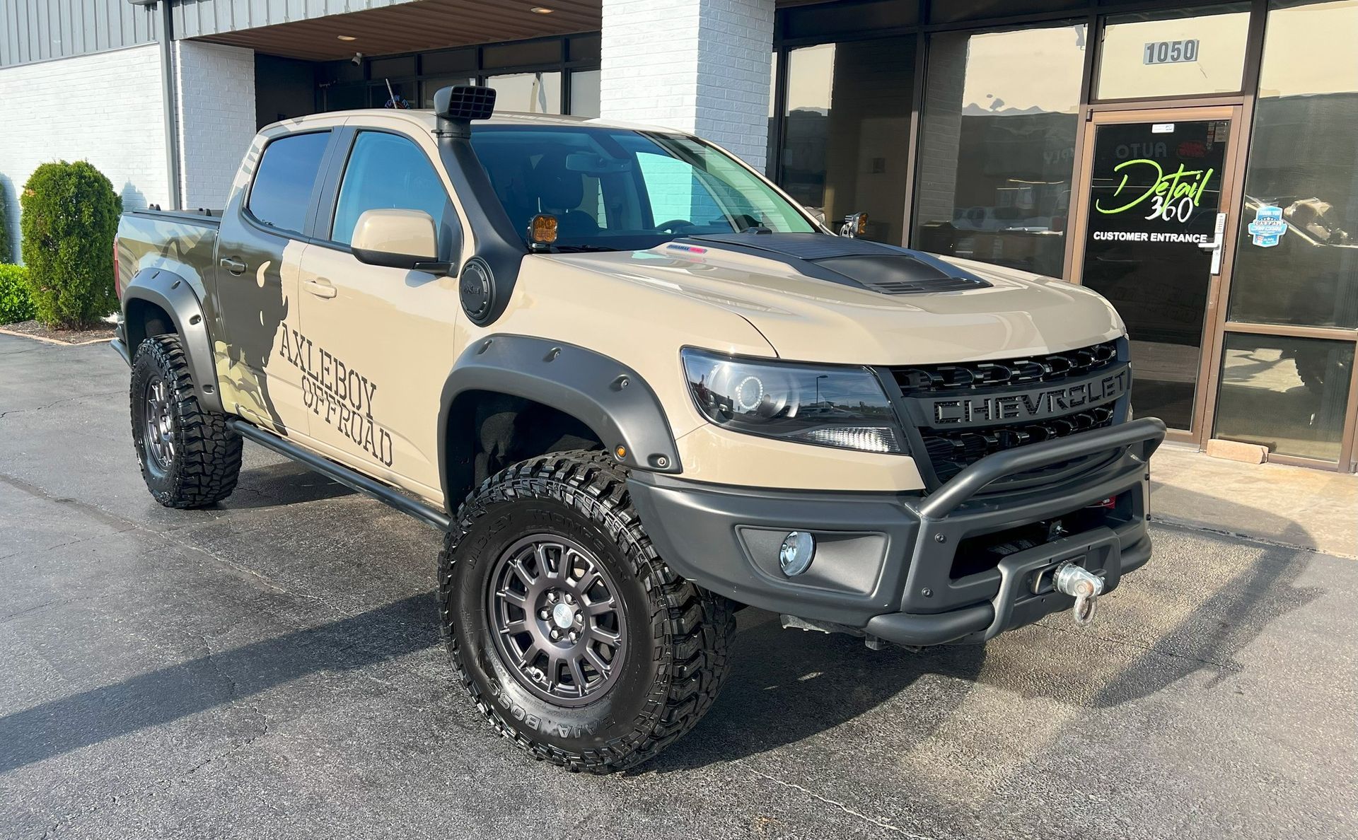 Tan Chevrolet Colorado truck with off-road modifications, parked outside a building.