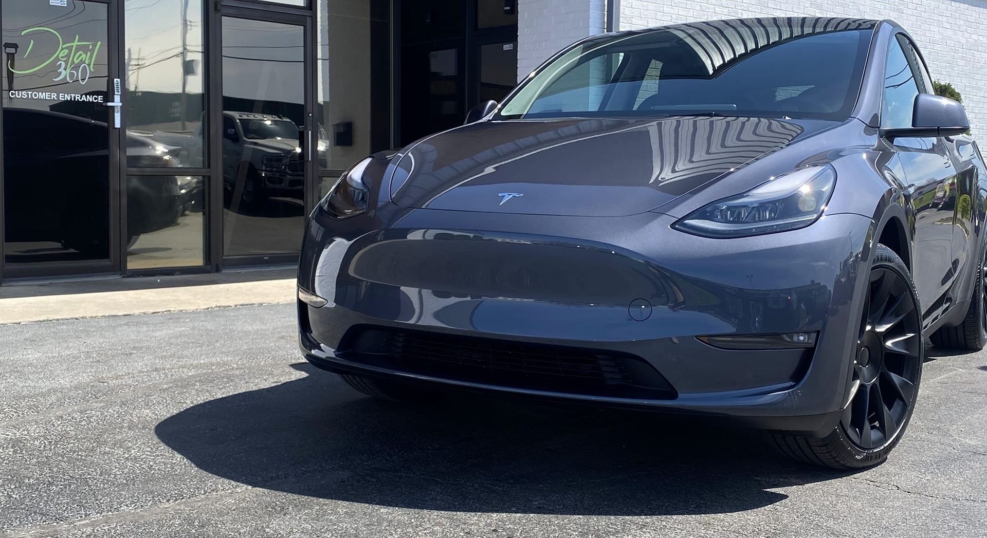 Gray Tesla Model Y parked in front of a building with black wheels and tinted windows on a sunny day.