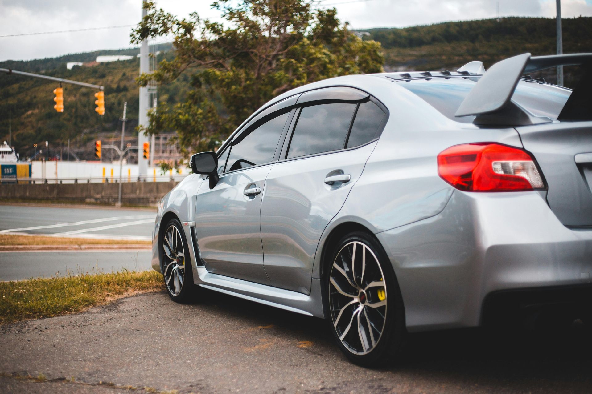 Silver Subaru WRX sedan parked on a roadside with a rear spoiler, dark tinted windows, and black and yellow rims.