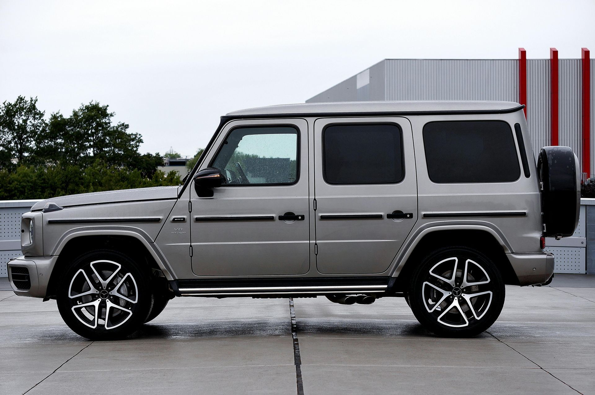 Silver Mercedes-Benz G-Class SUV parked on a gray surface with trees and a building in the background.
