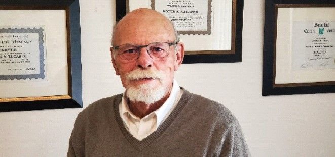 A man with a beard and glasses is standing in front of a wall with pictures on it.