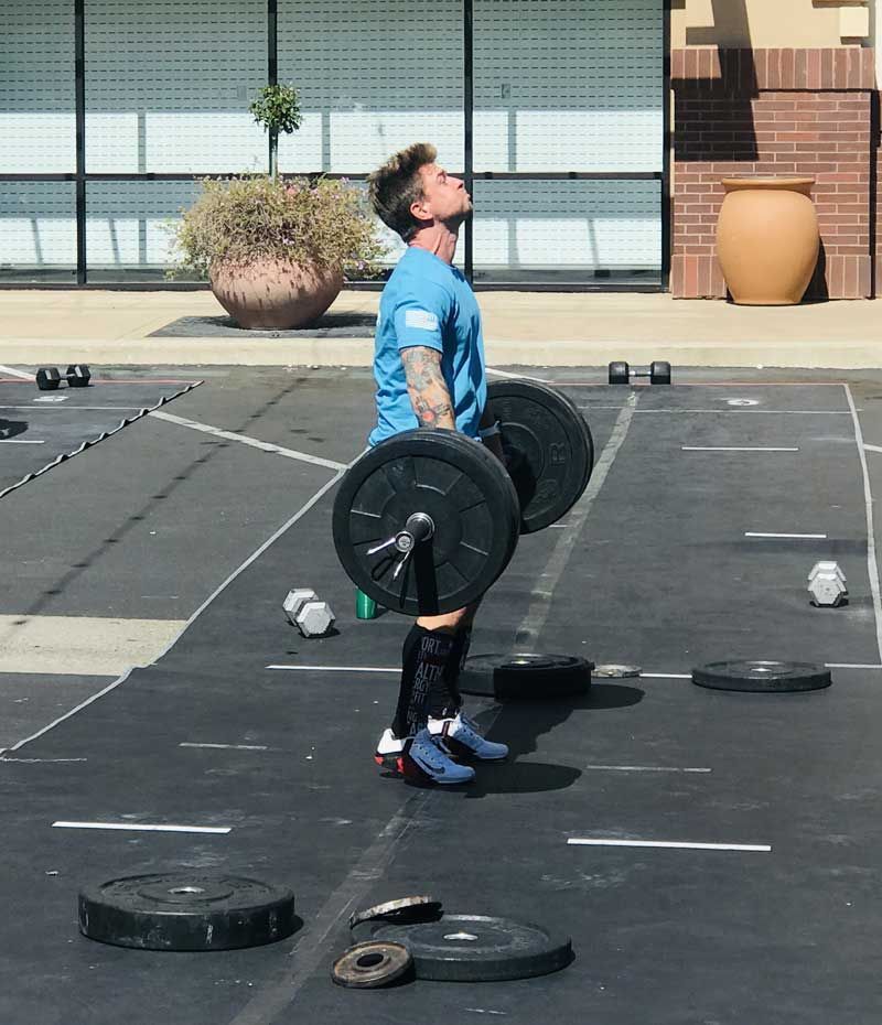 Person lifting a barbell outdoors, looking up. Wearing a blue shirt, compression socks, and sneakers.