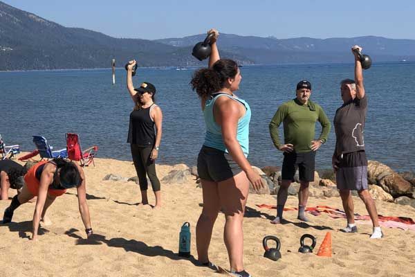 People exercising with kettlebells on a sunny beach on South Lake Tahoe, mountains in the background.