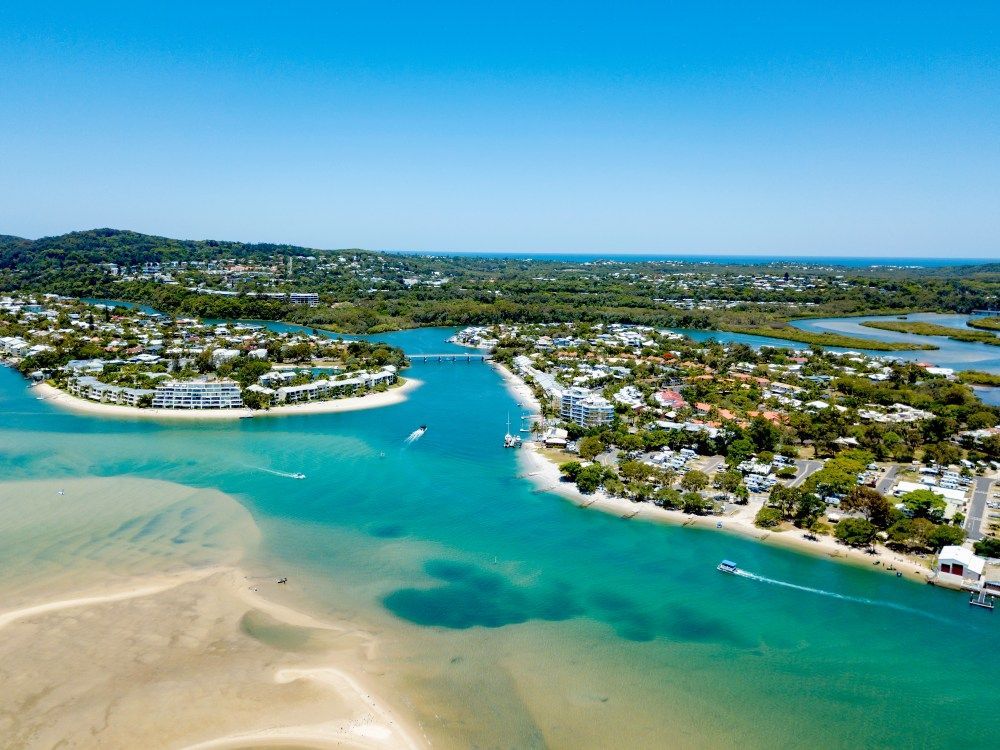 Aerial view of Noosa Heads, Australia, with blue water, sandy shores, buildings, and boats under a sunny sky.