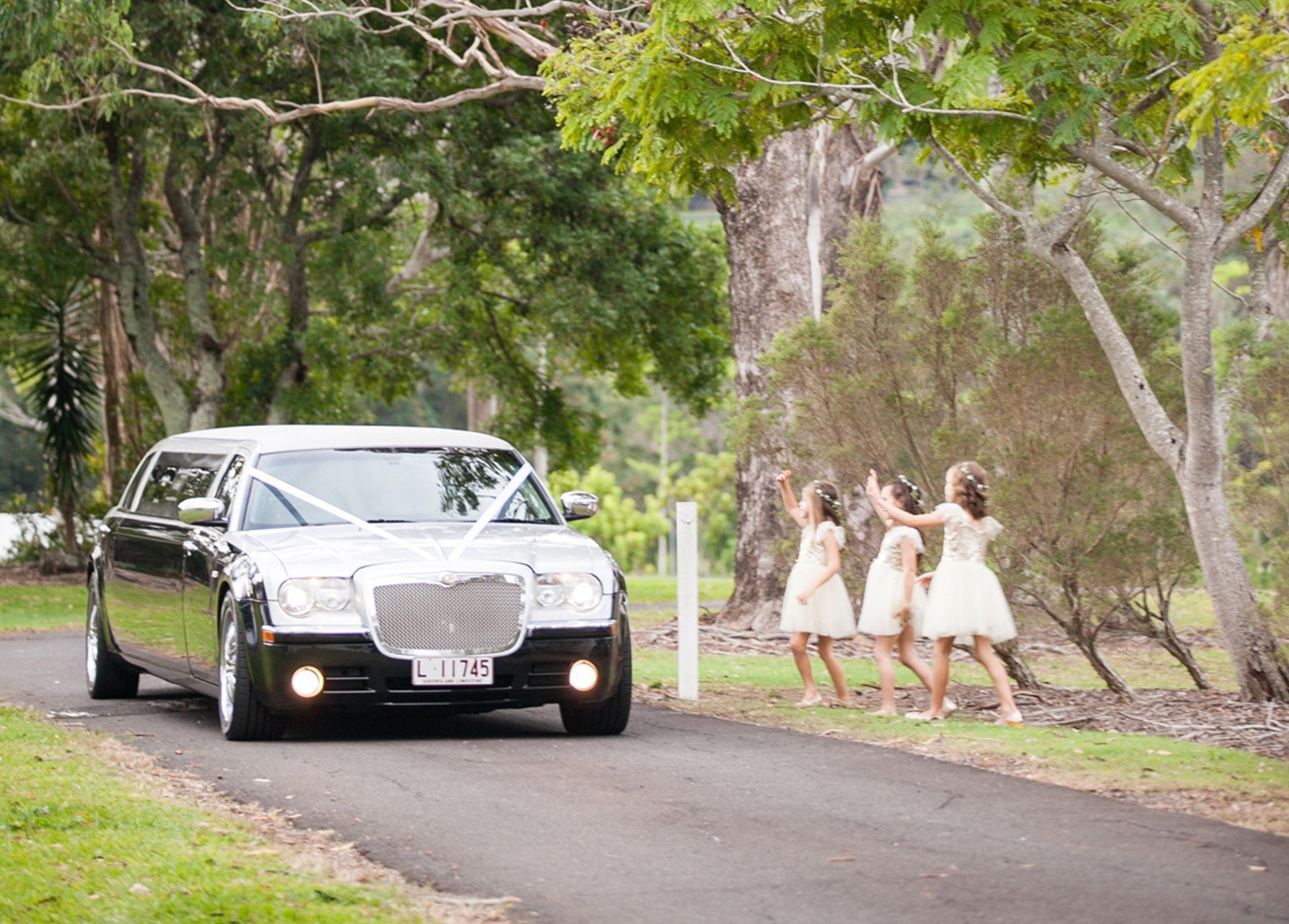 Black Limousine Drives Down a Road, Approaching Three Young Girls in Light Dresses Throwing Confetti — Superstretch300 Limousines in Sunshine Coast, QLD