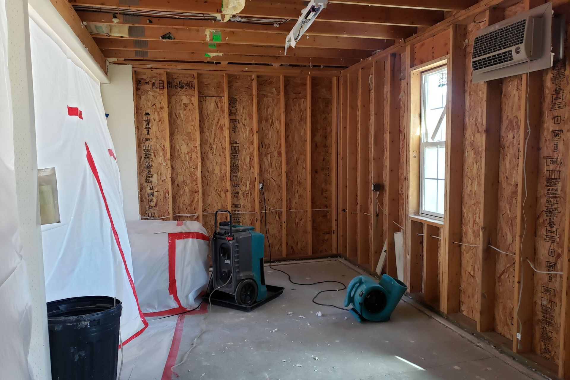 Interior room with exposed studs, equipment for remediation, plastic sheeting, and a small window.