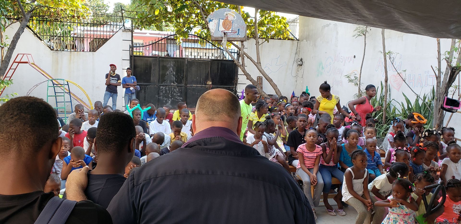 Man speaking into microphone, arm raised, addressing a crowd of children outdoors.