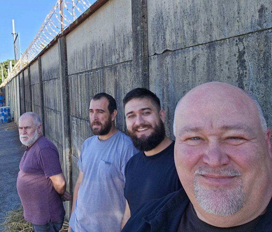 Four men pose in front of a concrete wall topped with barbed wire; the man on the right smiles.