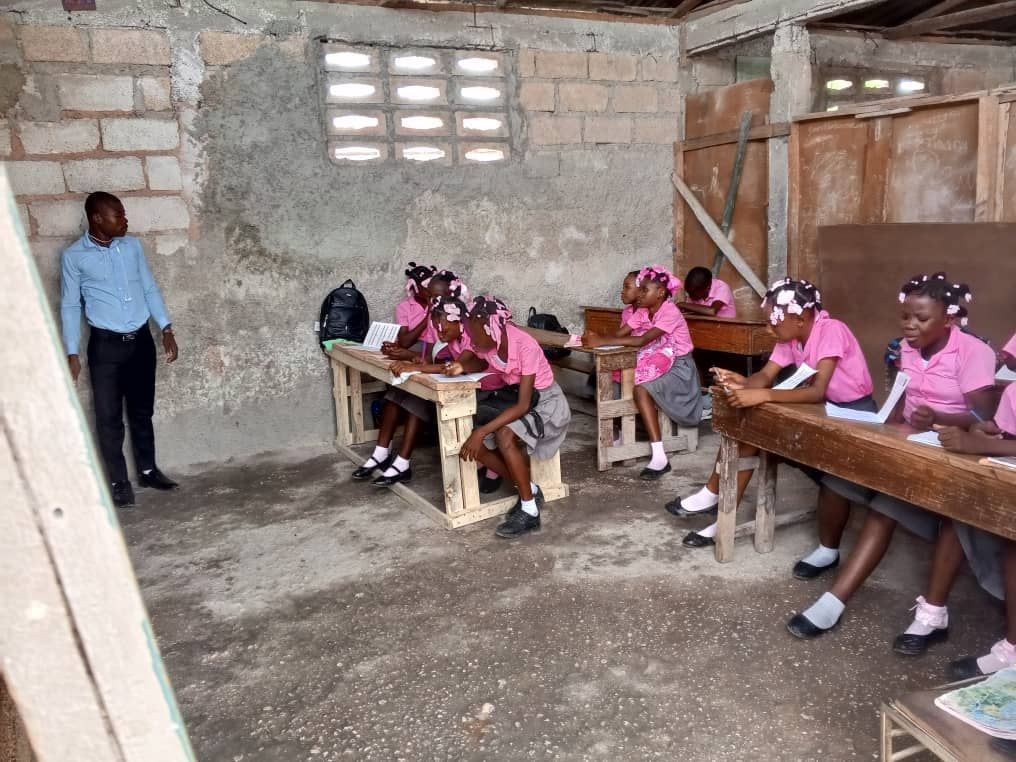 Teacher in blue shirt stands near students in pink uniforms writing at desks in a simple classroom.