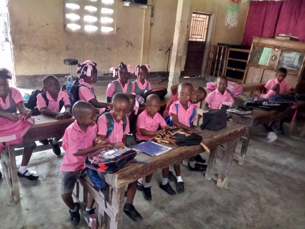 Children in pink shirts and skirts sit at desks in a classroom.